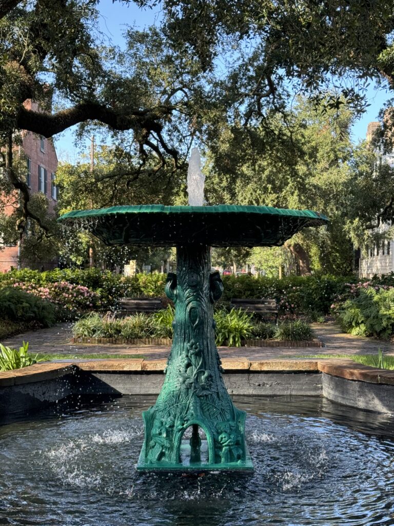 Green fountain in a park surrounded by oak trees and benches.