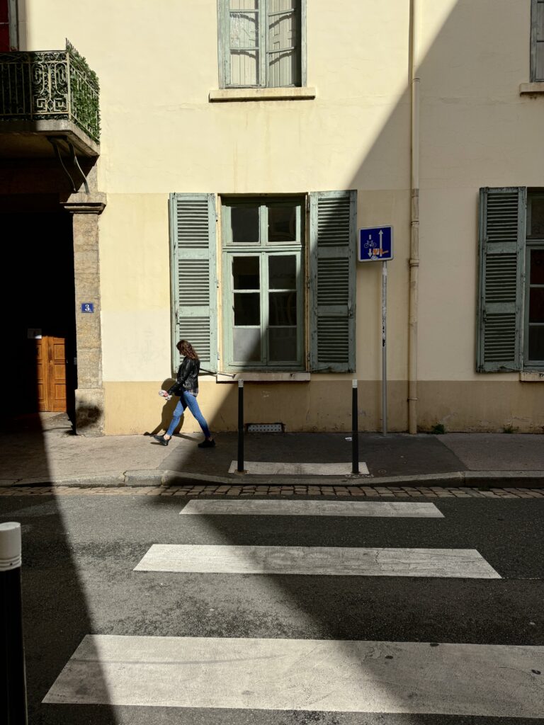 Woman walking on the street by the pedestrian crosswalk. She's walking in front of a large window with green shutters. 