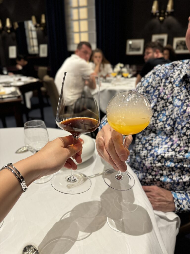 Two people toasting with wine glasses. One of them is filled with red wine and one of them with an orange liquid. 