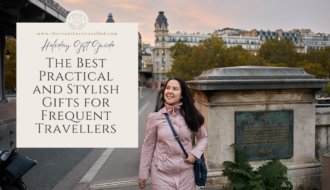 A woman wearing a vibrant pink coat and carrying a stylish blue bag strolls along a cobblestone street in Paris. The scene features iconic Parisian architecture in the background, with ornate balconies and decorative ironwork. She appears to be enjoying the crisp weather, her hair slightly tousled by the breeze, as she walks past charming cafes and boutique storefronts.