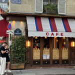 Street view of a Parisian café with a red, white, and blue striped awning labeled 'Café.' The exterior features wooden details, warm yellow lighting, and decorative patterns on the lower window panels. A green plant climbs along the wall under a sign for 'Rue Bonaparte.' Two people stand in the foreground looking at a phone, and outdoor seating with people dining is visible on the left.