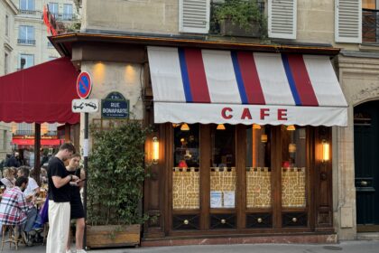 Street view of a Parisian café with a red, white, and blue striped awning labeled 'Café.' The exterior features wooden details, warm yellow lighting, and decorative patterns on the lower window panels. A green plant climbs along the wall under a sign for 'Rue Bonaparte.' Two people stand in the foreground looking at a phone, and outdoor seating with people dining is visible on the left.