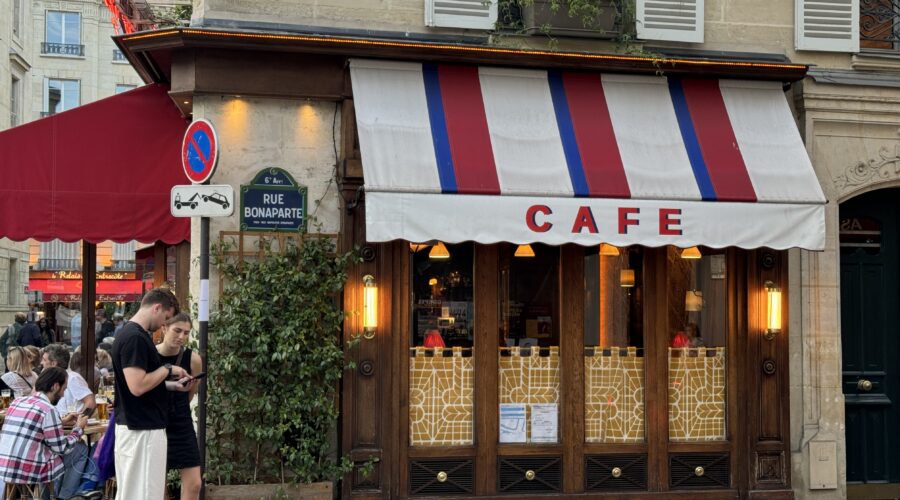 Street view of a Parisian café with a red, white, and blue striped awning labeled 'Café.' The exterior features wooden details, warm yellow lighting, and decorative patterns on the lower window panels. A green plant climbs along the wall under a sign for 'Rue Bonaparte.' Two people stand in the foreground looking at a phone, and outdoor seating with people dining is visible on the left.