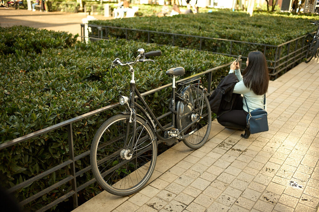 A woman wearing a light blue sweater and black leggings crouches down near a black bicycle, taking a photo with her smartphone. She carries a stylish blue crossbody travel bag, which holds her essentials securely. The sun casts a warm glow on the paved walkway, lined with neatly trimmed green bushes. Having a well-organized travel bag can tame pre-travel anxiety, keeping important items like passports, tickets, and phones within easy reach, so you can focus on capturing beautiful moments stress-free.