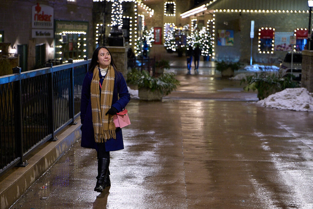 A woman in a navy blue coat, plaid scarf, and black boots walks confidently through a beautifully lit shopping district on a winter evening. She carries a pink purse and smiles as she enjoys the peaceful atmosphere, surrounded by festive string lights reflecting off the wet pavement. Overcoming pre-travel anxiety and travel-related stress, she looks relaxed and ready for her next adventure. Mastering pre-travel jitters can turn your trip into a walk in the park—just like this moment of calm before an exciting journey.