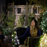 A woman in a navy blue coat and plaid scarf sits in a sleigh by a warmly lit shop setting, gazing up at the twinkling holiday lights. Surrounded by decorated Christmas trees and greenery, she appears calm and excited for her next adventure. Having a great travel bag can tame pre-travel anxiety, keeping essentials secure so you can enjoy magical moments like this without stress. With the right preparation, overcoming travel jitters turns every trip into a smooth and joyful experience.