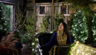 A woman in a navy blue coat and plaid scarf sits in a sleigh by a warmly lit shop setting, gazing up at the twinkling holiday lights. Surrounded by decorated Christmas trees and greenery, she appears calm and excited for her next adventure. Having a great travel bag can tame pre-travel anxiety, keeping essentials secure so you can enjoy magical moments like this without stress. With the right preparation, overcoming travel jitters turns every trip into a smooth and joyful experience.